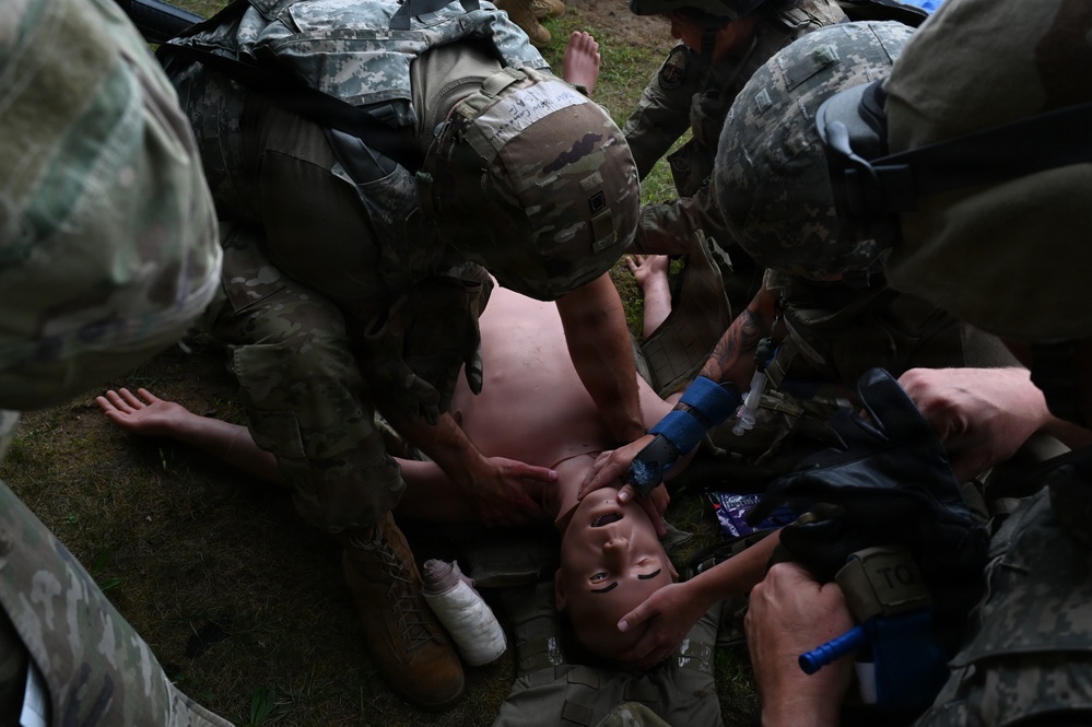 Multiple soldiers in US Army ACU/OCP camouflage uniforms are performing tactical combat casualty care (TCCC) on a shirtless simulated casualty lying on the ground. The scene appears to be a field medical training exercise, with personnel applying what appears to be a tourniquet and airway management techniques. Several soldiers are clustered around the patient simultaneously performing different medical interventions.