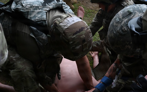 Multiple soldiers in US Army ACU/OCP camouflage uniforms are performing tactical combat casualty care (TCCC) on a shirtless simulated casualty lying on the ground. The scene appears to be a field medical training exercise, with personnel applying what appears to be a tourniquet and airway management techniques. Several soldiers are clustered around the patient simultaneously performing different medical interventions.