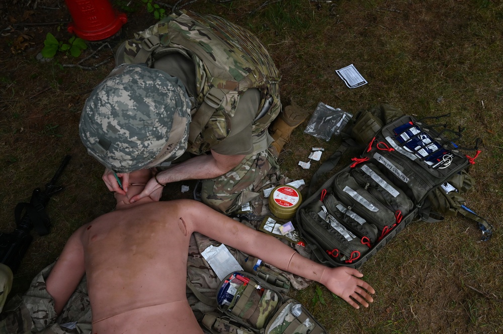 A military medic in camouflage uniform is performing a tactical combat casualty care (TCCC) training exercise on a medical mannequin. The scene includes an open medical aid bag with multiple supplies scattered on the ground. This appears to be a field training exercise based on the outdoor setting and training equipment present.