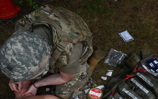 A military medic in camouflage uniform is performing a tactical combat casualty care (TCCC) training exercise on a medical mannequin. The scene includes an open medical aid bag with multiple supplies scattered on the ground. This appears to be a field training exercise based on the outdoor setting and training equipment present.