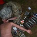 A military medic in camouflage uniform is performing a tactical combat casualty care (TCCC) training exercise on a medical mannequin. The scene includes an open medical aid bag with multiple supplies scattered on the ground. This appears to be a field training exercise based on the outdoor setting and training equipment present.