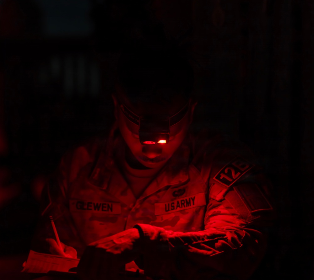A U.S. Army soldier identified by the name tape 'CLEWEN' is working in near-total darkness, illuminated solely by a red-light headlamp. The soldier appears to be writing or reviewing documents while wearing full combat uniform. The red lighting is consistent with tactical night operations, as red light preserves night vision.
