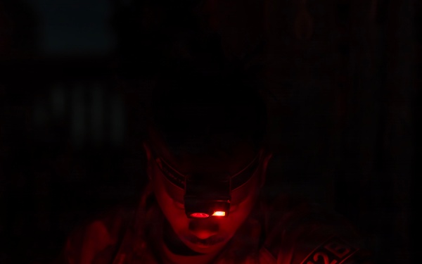 A U.S. Army soldier identified by the name tape 'CLEWEN' is working in near-total darkness, illuminated solely by a red-light headlamp. The soldier appears to be writing or reviewing documents while wearing full combat uniform. The red lighting is consistent with tactical night operations, as red light preserves night vision.