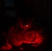 A U.S. Army soldier identified by the name tape 'CLEWEN' is working in near-total darkness, illuminated solely by a red-light headlamp. The soldier appears to be writing or reviewing documents while wearing full combat uniform. The red lighting is consistent with tactical night operations, as red light preserves night vision.