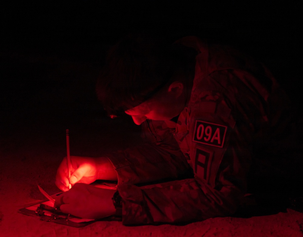 A military personnel wearing camouflage uniform is writing on a clipboard in near-total darkness, illuminated only by a red light source. The individual bears a visible patch marked '09A' on their sleeve, consistent with military identification markings. The use of red light is a tactical practice that preserves night vision while allowing task completion in low-light conditions.