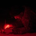 A military personnel wearing camouflage uniform is writing on a clipboard in near-total darkness, illuminated only by a red light source. The individual bears a visible patch marked '09A' on their sleeve, consistent with military identification markings. The use of red light is a tactical practice that preserves night vision while allowing task completion in low-light conditions.