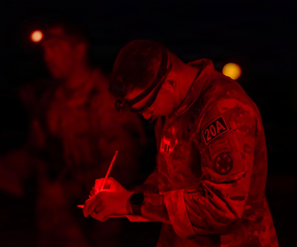 A military soldier is writing on a document or clipboard at night, illuminated by red light. The soldier is wearing camouflage uniform with a '20A' patch visible on the shoulder, along with a headlamp. Additional personnel are visible in the blurred background, also under red lighting conditions.