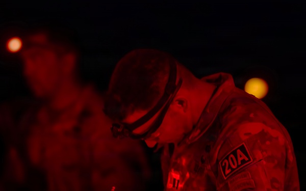 A military soldier is writing on a document or clipboard at night, illuminated by red light. The soldier is wearing camouflage uniform with a '20A' patch visible on the shoulder, along with a headlamp. Additional personnel are visible in the blurred background, also under red lighting conditions.