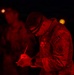 A military soldier is writing on a document or clipboard at night, illuminated by red light. The soldier is wearing camouflage uniform with a '20A' patch visible on the shoulder, along with a headlamp. Additional personnel are visible in the blurred background, also under red lighting conditions.