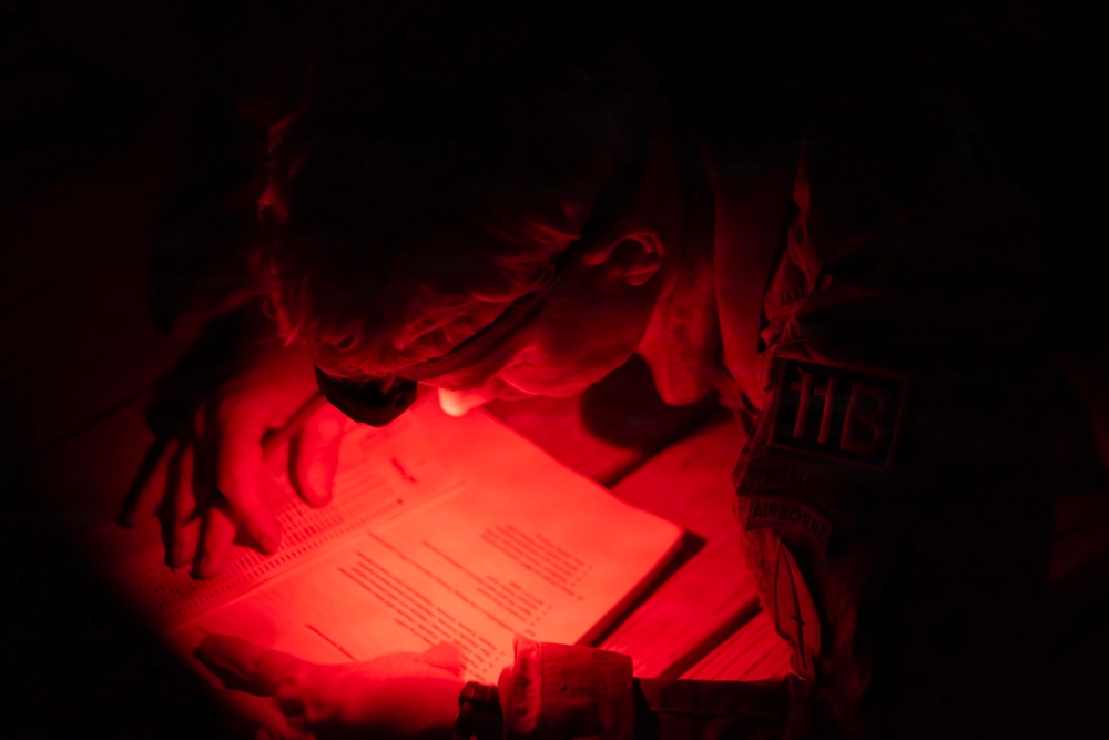 A person, appearing to be in military or tactical attire, is reading or reviewing documents under red light in a dark environment. The red light illumination is consistent with night operations, as red light preserves night vision. A patch or insignia with the number '113' is visible on their shoulder.