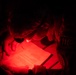 A person, appearing to be in military or tactical attire, is reading or reviewing documents under red light in a dark environment. The red light illumination is consistent with night operations, as red light preserves night vision. A patch or insignia with the number '113' is visible on their shoulder.