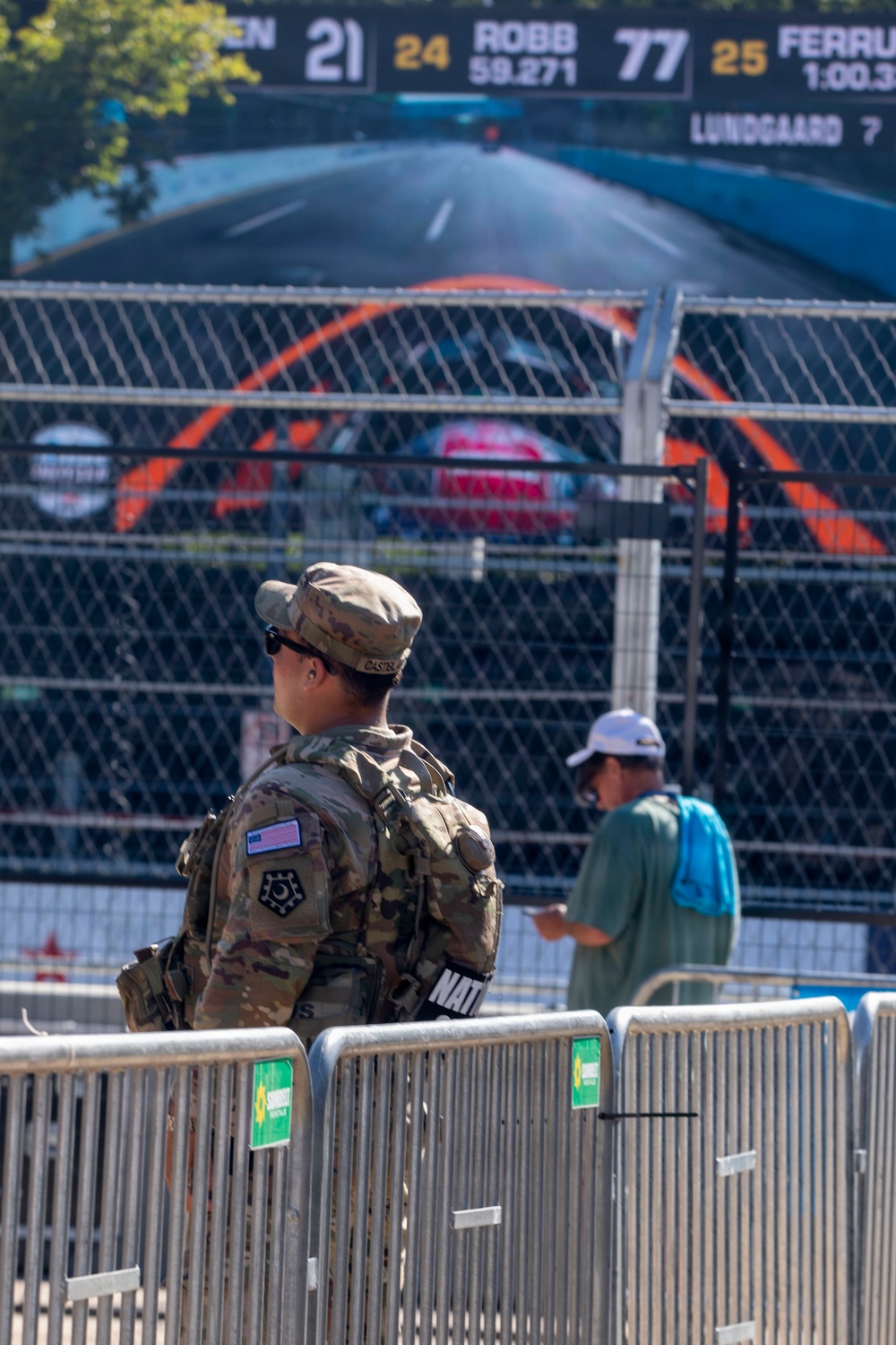 A U.S. Army soldier in MultiCam camouflage uniform with an American flag patch and 'NAT' (National Guard) armband stands at a security barrier at what appears to be a motorsport racing event. A civilian in a teal shirt and white cap with a blue backpack stands nearby, looking at a phone. A large scoreboard in the background displays driver names and lap times including ROBB (24, 59.271), number 77, FERRU (25, 1:00.3), and LUNDGAARD (7).