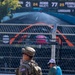 A U.S. Army soldier in MultiCam camouflage uniform with an American flag patch and 'NAT' (National Guard) armband stands at a security barrier at what appears to be a motorsport racing event. A civilian in a teal shirt and white cap with a blue backpack stands nearby, looking at a phone. A large scoreboard in the background displays driver names and lap times including ROBB (24, 59.271), number 77, FERRU (25, 1:00.3), and LUNDGAARD (7).