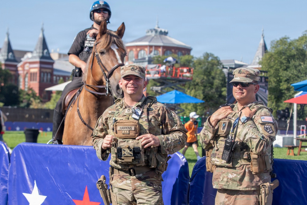 Two U.S. Army soldiers in MultiCam (OCP) camouflage uniforms and tactical vests are standing at what appears to be an outdoor public event, likely on the National Mall in Washington, D.C., based on the Smithsonian Institution Building visible in the background. A mounted park police officer on a brown horse is positioned behind them. The scene suggests a security or ceremonial presence at a large public gathering.