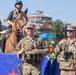 Two U.S. Army soldiers in MultiCam (OCP) camouflage uniforms and tactical vests are standing at what appears to be an outdoor public event, likely on the National Mall in Washington, D.C., based on the Smithsonian Institution Building visible in the background. A mounted park police officer on a brown horse is positioned behind them. The scene suggests a security or ceremonial presence at a large public gathering.