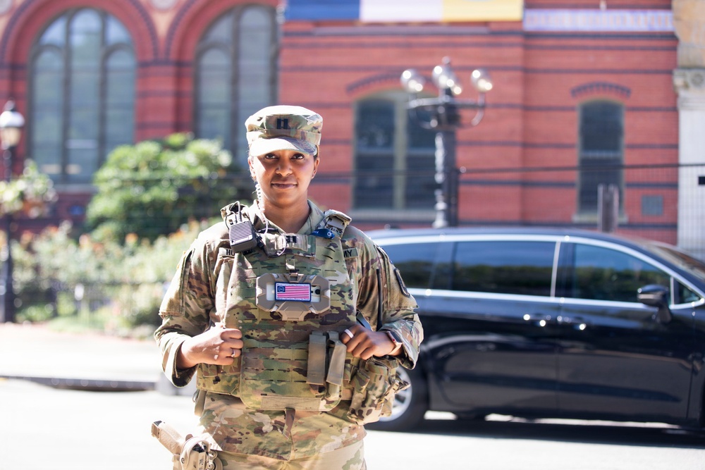 A female U.S. Army soldier in full Operational Camouflage Pattern (OCP) uniform stands outdoors in front of a red brick building. She is wearing a tactical plate carrier vest with a U.S. flag patch and a captain's rank insignia on her patrol cap. The setting appears to be an urban environment, possibly Washington D.C., based on the architectural style.