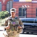 A female U.S. Army soldier in full Operational Camouflage Pattern (OCP) uniform stands outdoors in front of a red brick building. She is wearing a tactical plate carrier vest with a U.S. flag patch and a captain's rank insignia on her patrol cap. The setting appears to be an urban environment, possibly Washington D.C., based on the architectural style.
