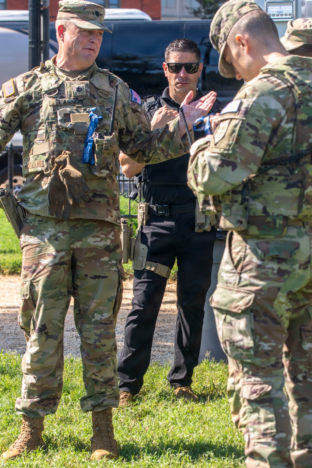 Three individuals are engaged in a conversation outdoors on a grassy area. Two are wearing U.S. Army OCP (Operational Camouflage Pattern) uniforms with tactical gear, while a third individual wears black tactical clothing with a holstered sidearm. The leftmost soldier has a visible name tape reading 'CANNON' and wears a rank insignia consistent with a Lieutenant Colonel (oak leaf cluster).