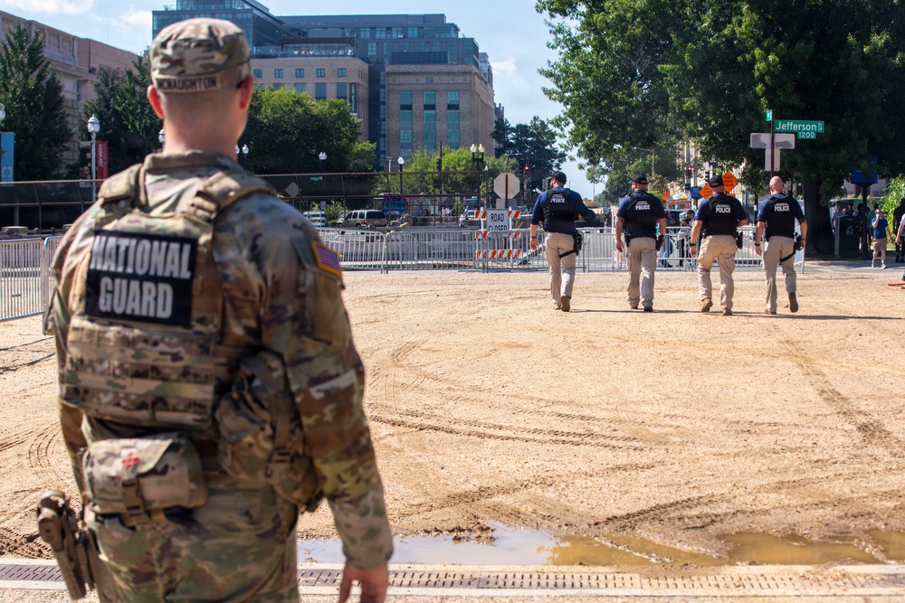 A National Guard soldier stands in the foreground observing a group of police officers walking across a cleared sandy/dirt area. The scene appears to be a security deployment at what looks like a controlled perimeter zone in an urban setting. A street sign for Jefferson Street is visible in the background along with barriers and urban buildings.