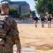 A National Guard soldier stands in the foreground observing a group of police officers walking across a cleared sandy/dirt area. The scene appears to be a security deployment at what looks like a controlled perimeter zone in an urban setting. A street sign for Jefferson Street is visible in the background along with barriers and urban buildings.