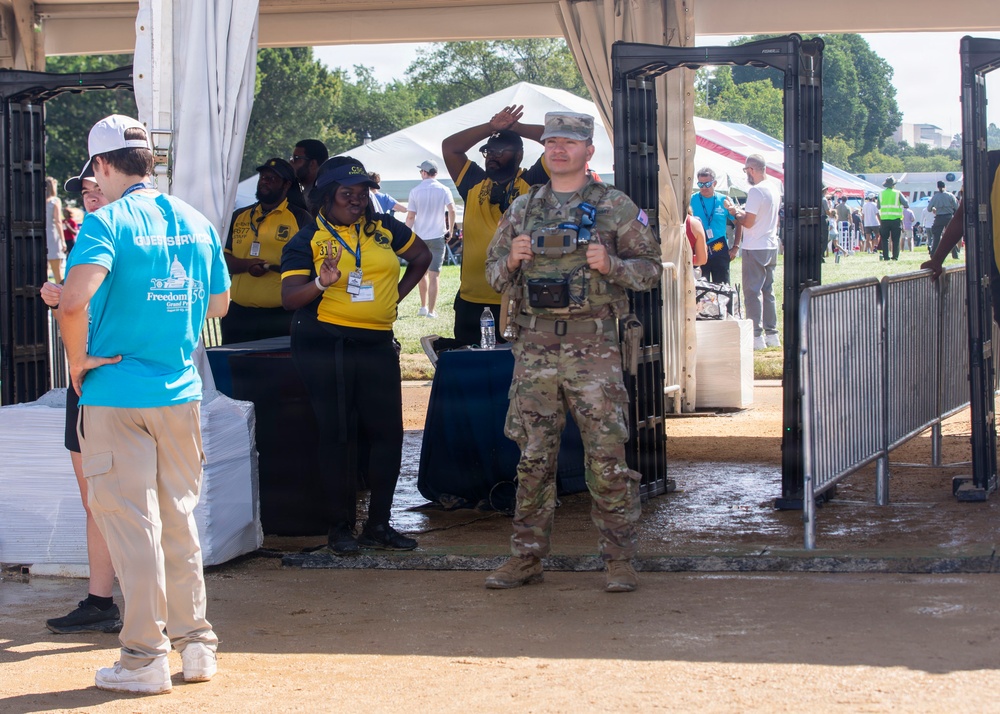The image shows a security checkpoint at an outdoor public event, likely on the National Mall in Washington D.C., based on visible signage referencing 'Freedom' and the Capitol dome logo. A uniformed military soldier in multicam/OCP camouflage stands at a metal detector portal alongside civilian security personnel wearing yellow polo shirts. Event volunteers in light blue shirts are also present near the entry point.