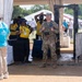 The image shows a security checkpoint at an outdoor public event, likely on the National Mall in Washington D.C., based on visible signage referencing 'Freedom' and the Capitol dome logo. A uniformed military soldier in multicam/OCP camouflage stands at a metal detector portal alongside civilian security personnel wearing yellow polo shirts. Event volunteers in light blue shirts are also present near the entry point.
