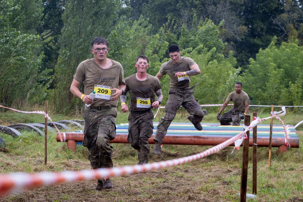 A group of participants are competing in a military-style obstacle course race, navigating over large pipes and barriers on muddy terrain. The competitors are wearing military-style clothing and are visibly covered in mud, indicating they have already passed through several obstacles. Race bibs with numbers 209, 295, and 057 are visible on the front runners.