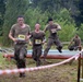 A group of participants are competing in a military-style obstacle course race, navigating over large pipes and barriers on muddy terrain. The competitors are wearing military-style clothing and are visibly covered in mud, indicating they have already passed through several obstacles. Race bibs with numbers 209, 295, and 057 are visible on the front runners.