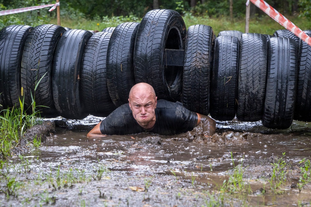 A bald man in a black shirt is crawling through muddy water beneath a barrier made of stacked tires during what appears to be an obstacle course race. His face and clothing are covered in mud, and he displays a determined expression. The setting is an outdoor muddy terrain with trees and caution tape visible in the background.
