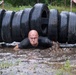 A bald man in a black shirt is crawling through muddy water beneath a barrier made of stacked tires during what appears to be an obstacle course race. His face and clothing are covered in mud, and he displays a determined expression. The setting is an outdoor muddy terrain with trees and caution tape visible in the background.