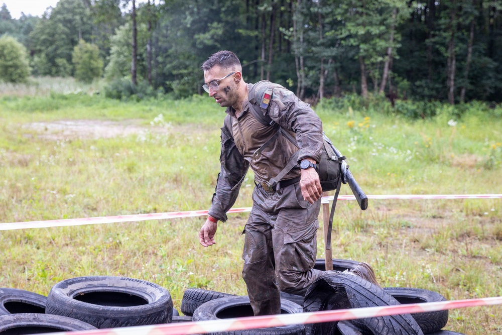 A mud-covered soldier or military competitor navigates a tire obstacle course in an outdoor field setting. The individual is wearing military-style fatigues heavily soiled with mud, safety glasses, and a watch, suggesting participation in a military fitness or obstacle race competition. Red and white barrier tape marks the course boundaries, with discarded tires scattered on the ground.