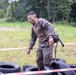 A mud-covered soldier or military competitor navigates a tire obstacle course in an outdoor field setting. The individual is wearing military-style fatigues heavily soiled with mud, safety glasses, and a watch, suggesting participation in a military fitness or obstacle race competition. Red and white barrier tape marks the course boundaries, with discarded tires scattered on the ground.