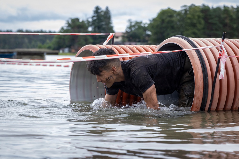 A man is crawling through a large corrugated plastic pipe tunnel that is partially submerged in water, appearing to be part of an obstacle course race. He is wearing a black t-shirt and is visibly wet and muddy as he emerges from the pipe opening. The setting appears to be a lake or large body of water with trees and overcast skies in the background.