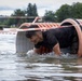 A man is crawling through a large corrugated plastic pipe tunnel that is partially submerged in water, appearing to be part of an obstacle course race. He is wearing a black t-shirt and is visibly wet and muddy as he emerges from the pipe opening. The setting appears to be a lake or large body of water with trees and overcast skies in the background.