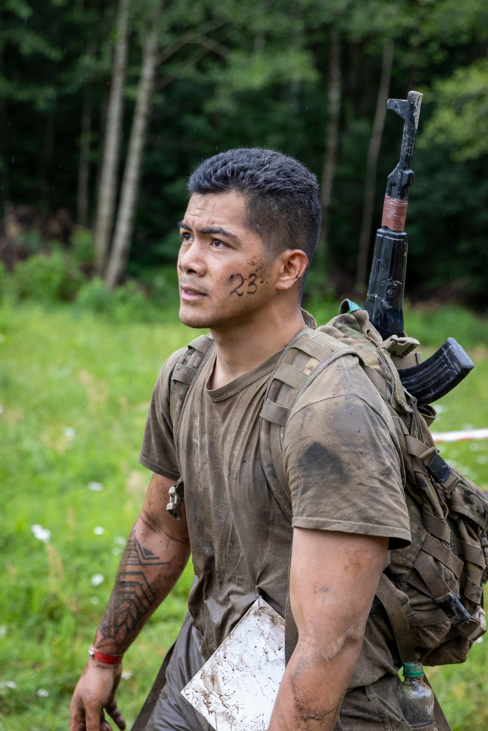 A young man in mud-covered military-style clothing carries a tactical backpack with a rifle (appearing to be an AK-pattern weapon) slung over his shoulder. The number '23' is written on his cheek, and he has visible tribal-style tattoos on his forearm. He is standing in an outdoor grassy area with a forested background, appearing to be participating in a military training exercise or competition.