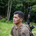 A young man in mud-covered military-style clothing carries a tactical backpack with a rifle (appearing to be an AK-pattern weapon) slung over his shoulder. The number '23' is written on his cheek, and he has visible tribal-style tattoos on his forearm. He is standing in an outdoor grassy area with a forested background, appearing to be participating in a military training exercise or competition.