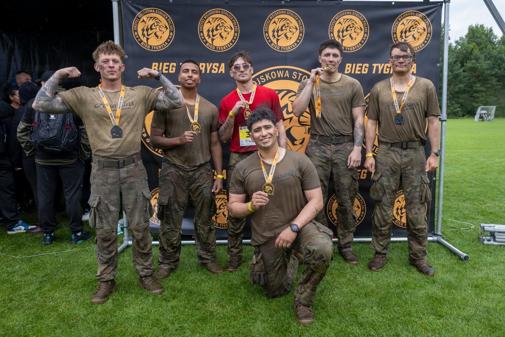 A group of six men pose for a photo in front of a 'Bieg Tygrysa' (Tiger Run) event banner in Poland. Most participants are wearing mud-covered military-style camouflage clothing and are displaying finisher medals. The group appears to be celebrating the completion of a military-themed obstacle/mud run event.