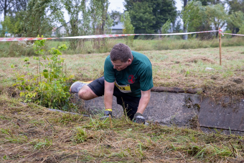A male participant is competing in an outdoor obstacle course race, crawling over a grass-covered terrain near a concrete barrier. He is wearing a green t-shirt, black shorts, gloves, and a yellow race bib numbered '13'. The course is marked with red and white barrier tape strung between poles in the background.