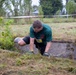 A male participant is competing in an outdoor obstacle course race, crawling over a grass-covered terrain near a concrete barrier. He is wearing a green t-shirt, black shorts, gloves, and a yellow race bib numbered '13'. The course is marked with red and white barrier tape strung between poles in the background.