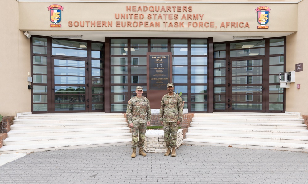 Two U.S. Army soldiers in OCP (Operational Camouflage Pattern) uniforms are posing for a photo in front of the Headquarters of the United States Army Southern European Task Force, Africa (SETAF-AF). The building entrance features a dedication plaque labeled 'Fox-Giunta Hall' and displays the SETAF-Africa unit patches on either side of the building facade. The setting appears to be a military installation, likely in Italy.