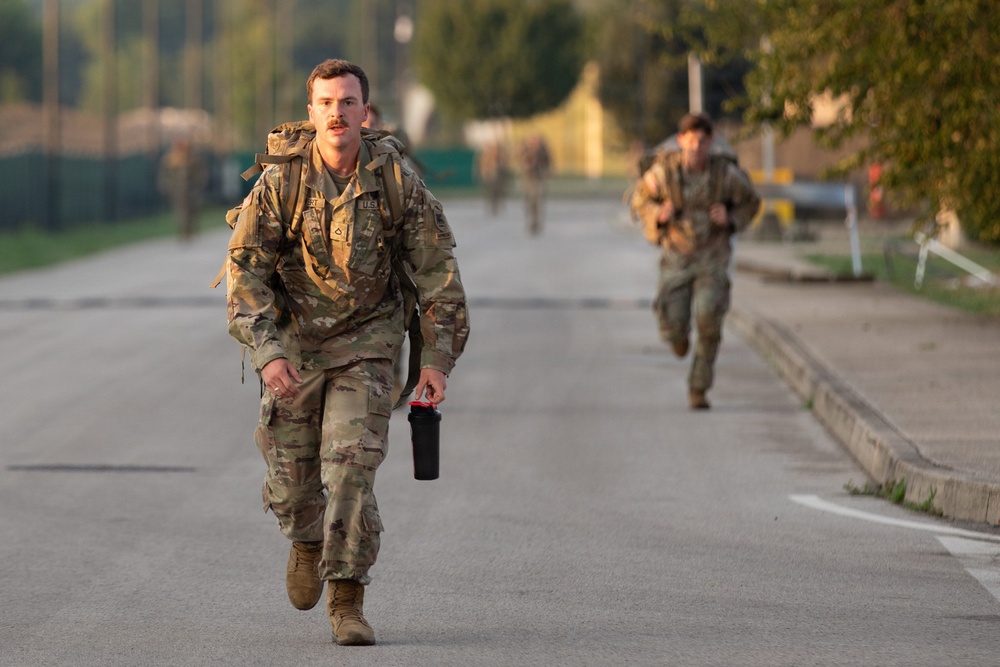 Multiple soldiers in US Army OCP (Operational Camouflage Pattern) uniforms are running along a paved road, likely during a physical training exercise or ruck march. The lead soldier in the foreground is carrying a large rucksack and a black water bottle, with additional soldiers visible in the background. The setting appears to be a military installation or training area during early morning or late afternoon light.