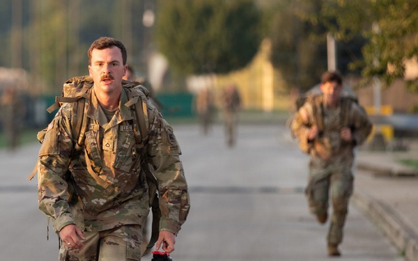 Multiple soldiers in US Army OCP (Operational Camouflage Pattern) uniforms are running along a paved road, likely during a physical training exercise or ruck march. The lead soldier in the foreground is carrying a large rucksack and a black water bottle, with additional soldiers visible in the background. The setting appears to be a military installation or training area during early morning or late afternoon light.