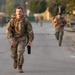 Multiple soldiers in US Army OCP (Operational Camouflage Pattern) uniforms are running along a paved road, likely during a physical training exercise or ruck march. The lead soldier in the foreground is carrying a large rucksack and a black water bottle, with additional soldiers visible in the background. The setting appears to be a military installation or training area during early morning or late afternoon light.