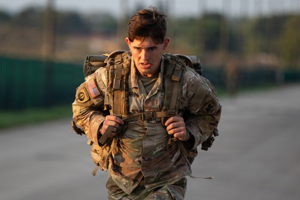 A young U.S. Army soldier in MultiCam (OCP) uniform is conducting a ruck march or physical training event, carrying a heavy military rucksack. The soldier displays a determined, strained expression suggesting significant physical exertion. The image was taken in warm, low-angle light consistent with early morning or late afternoon conditions.