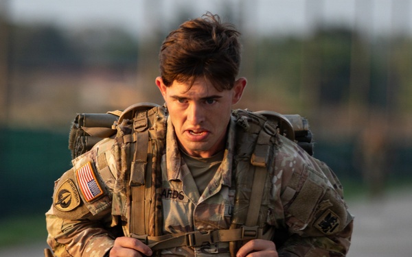 A young U.S. Army soldier in MultiCam (OCP) uniform is conducting a ruck march or physical training event, carrying a heavy military rucksack. The soldier displays a determined, strained expression suggesting significant physical exertion. The image was taken in warm, low-angle light consistent with early morning or late afternoon conditions.