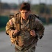 A young U.S. Army soldier in MultiCam (OCP) uniform is conducting a ruck march or physical training event, carrying a heavy military rucksack. The soldier displays a determined, strained expression suggesting significant physical exertion. The image was taken in warm, low-angle light consistent with early morning or late afternoon conditions.