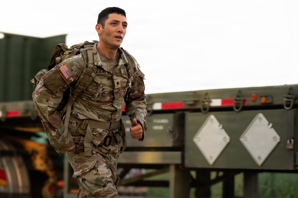A U.S. Army soldier in full OCP (Operational Camouflage Pattern) uniform is running while carrying a heavy rucksack. The soldier, identified by a name tape reading 'NICOLAS' and a U.S. Army tape, is moving past military vehicles including what appears to be a flatbed trailer. The setting appears to be an outdoor military training or operational environment.