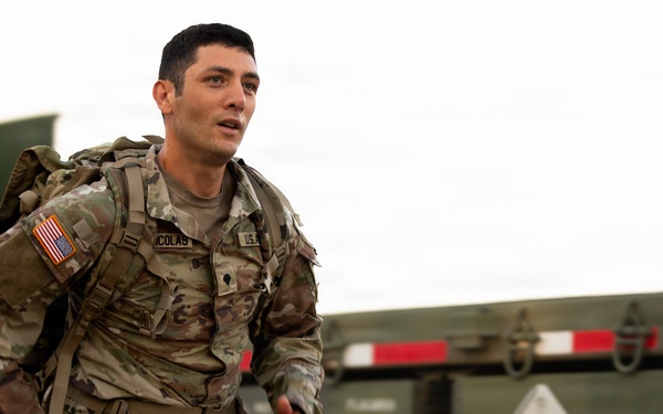 A U.S. Army soldier in full OCP (Operational Camouflage Pattern) uniform is running while carrying a heavy rucksack. The soldier, identified by a name tape reading 'NICOLAS' and a U.S. Army tape, is moving past military vehicles including what appears to be a flatbed trailer. The setting appears to be an outdoor military training or operational environment.
