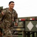 A U.S. Army soldier in full OCP (Operational Camouflage Pattern) uniform is running while carrying a heavy rucksack. The soldier, identified by a name tape reading 'NICOLAS' and a U.S. Army tape, is moving past military vehicles including what appears to be a flatbed trailer. The setting appears to be an outdoor military training or operational environment.