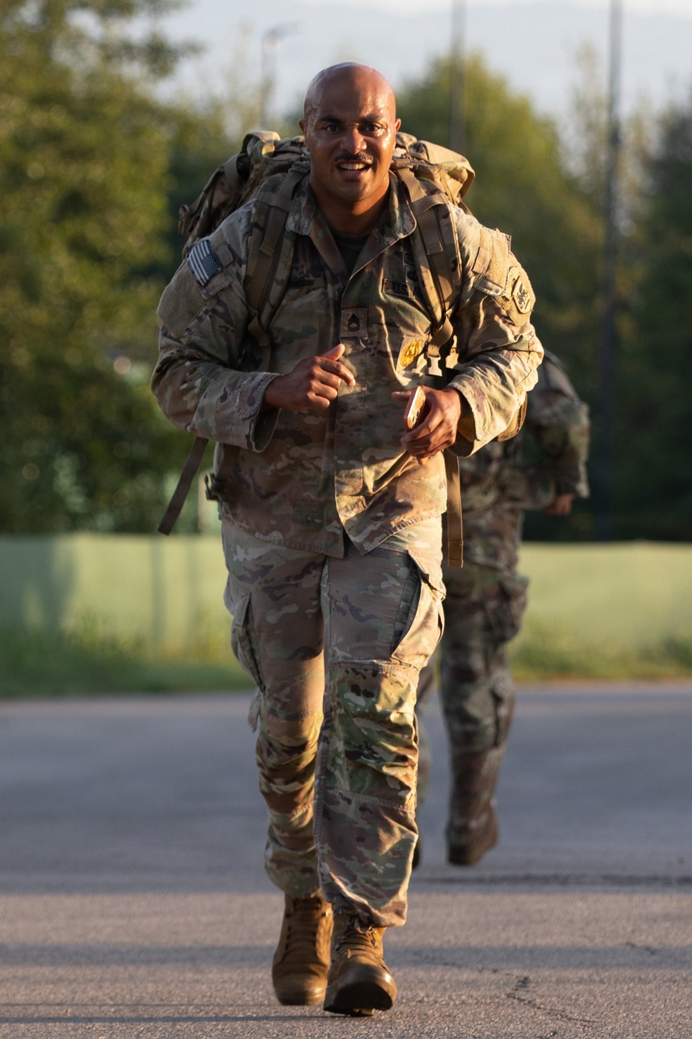A U.S. Army soldier in Operational Camouflage Pattern (OCP) uniform is running or rucking (ruck march) along a paved road while carrying a large military rucksack. The soldier is smiling and appears to be in good spirits despite the physical exertion. A second soldier in similar uniform is visible in the background, also carrying a rucksack.
